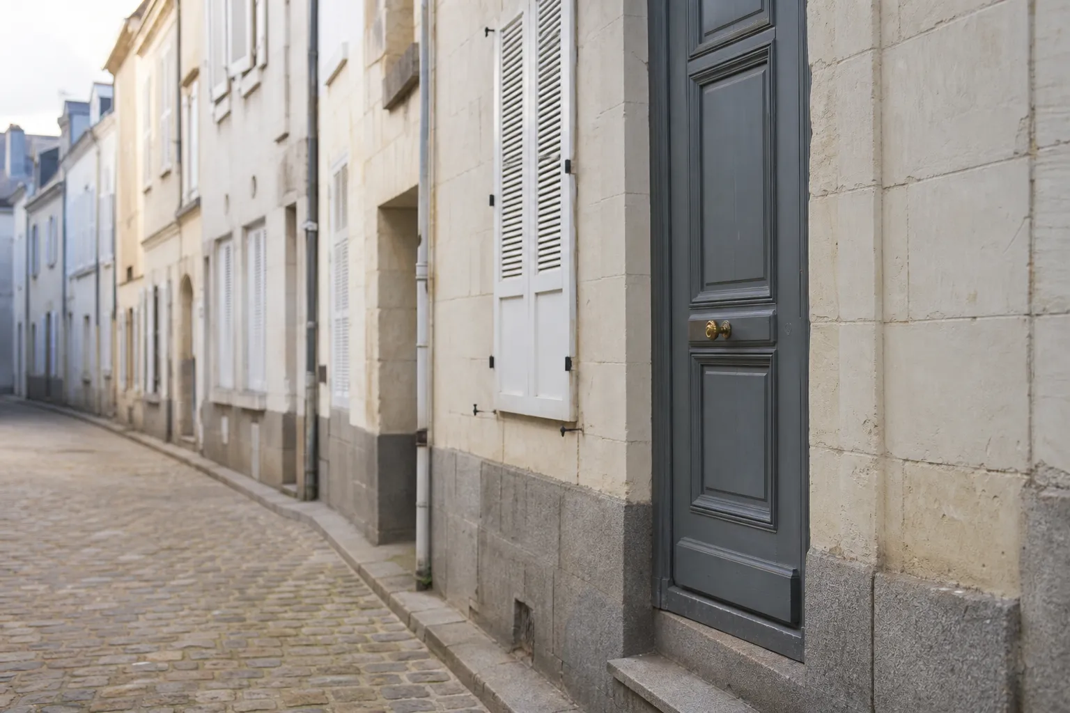 Façade en pierre de taille claire à soubassement de granit, volets de bois blancs, dans une rue pavée